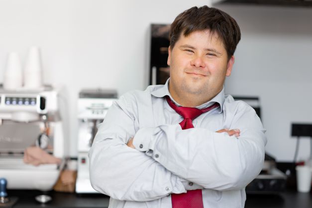 Young Down Syndrome man at work in cafe standing smiling with arms crossed, in front of coffee machine