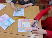 Table with artwork and arms of two participants creating their artwork with coloured pencils